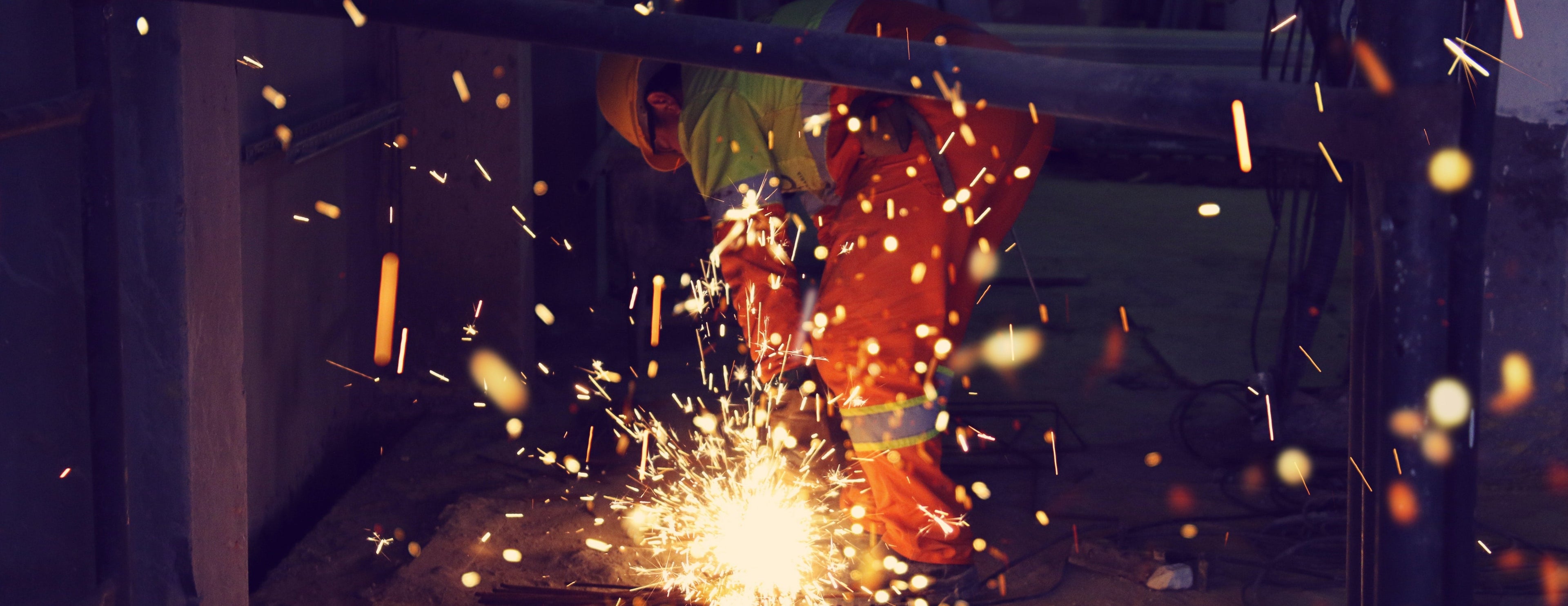 A man in an orange shirt engages in welding a metal piece, with sparks illuminating the workspace around him.